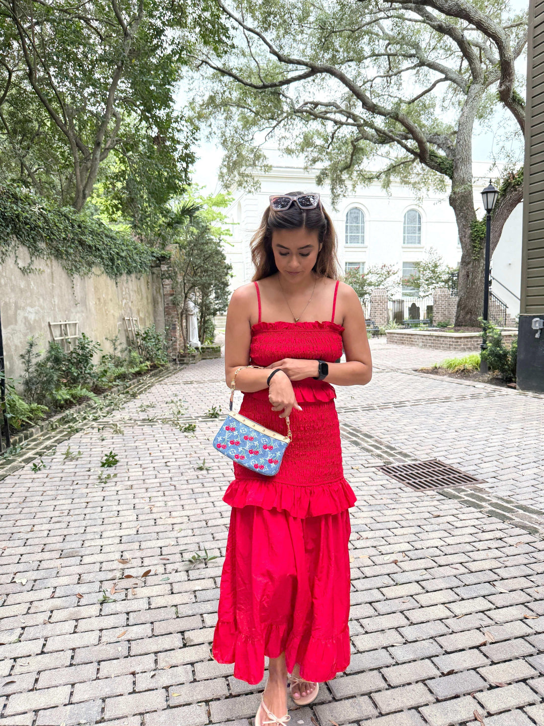Woman in a red dress holding a colorful handbag on a charming cobblestone street surrounded by greenery.