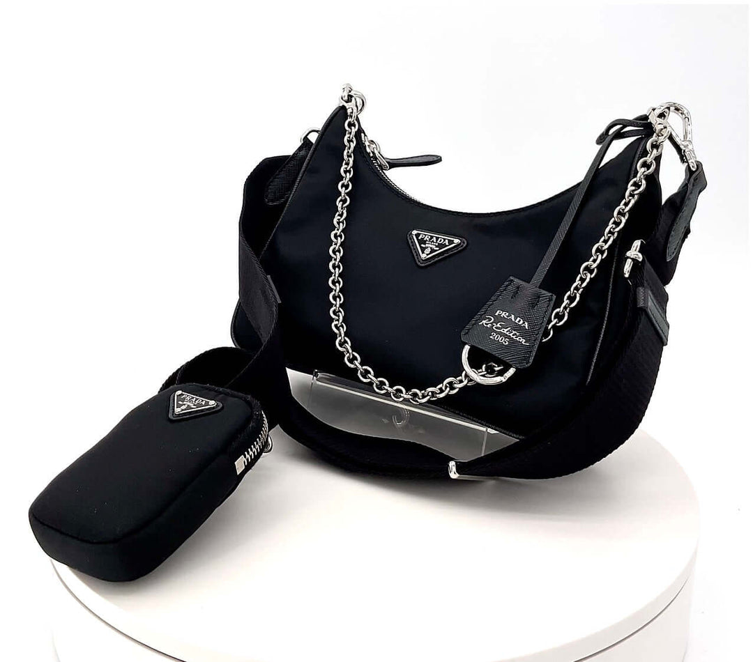 Authentic Prada handbag in black with Saffiano leather, silver-tone chain straps, and accessories on a white display.