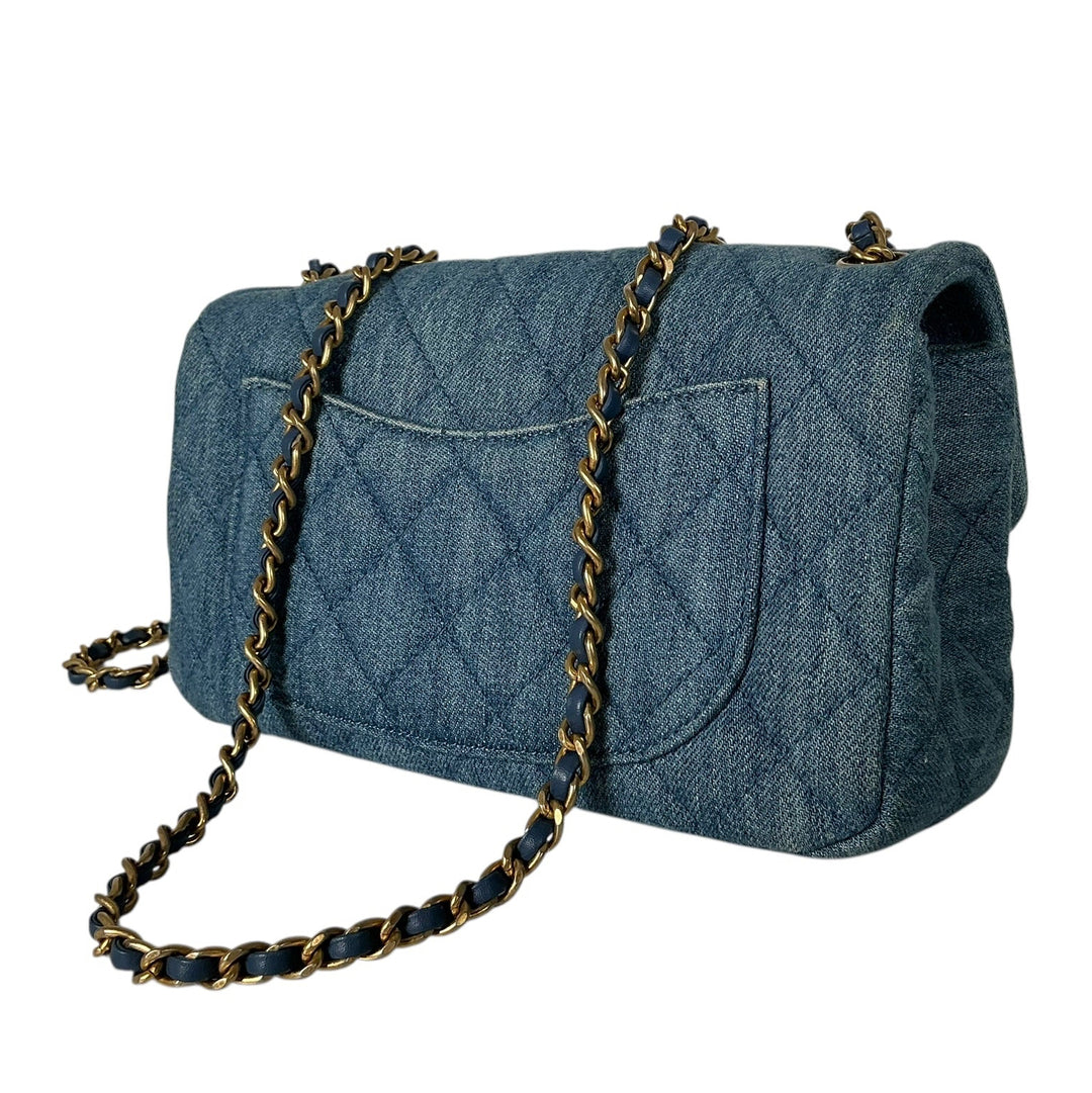 A quilted blue denim shoulder bag with a gold and black chain strap, shown from a three-quarters angle on a plain white background.