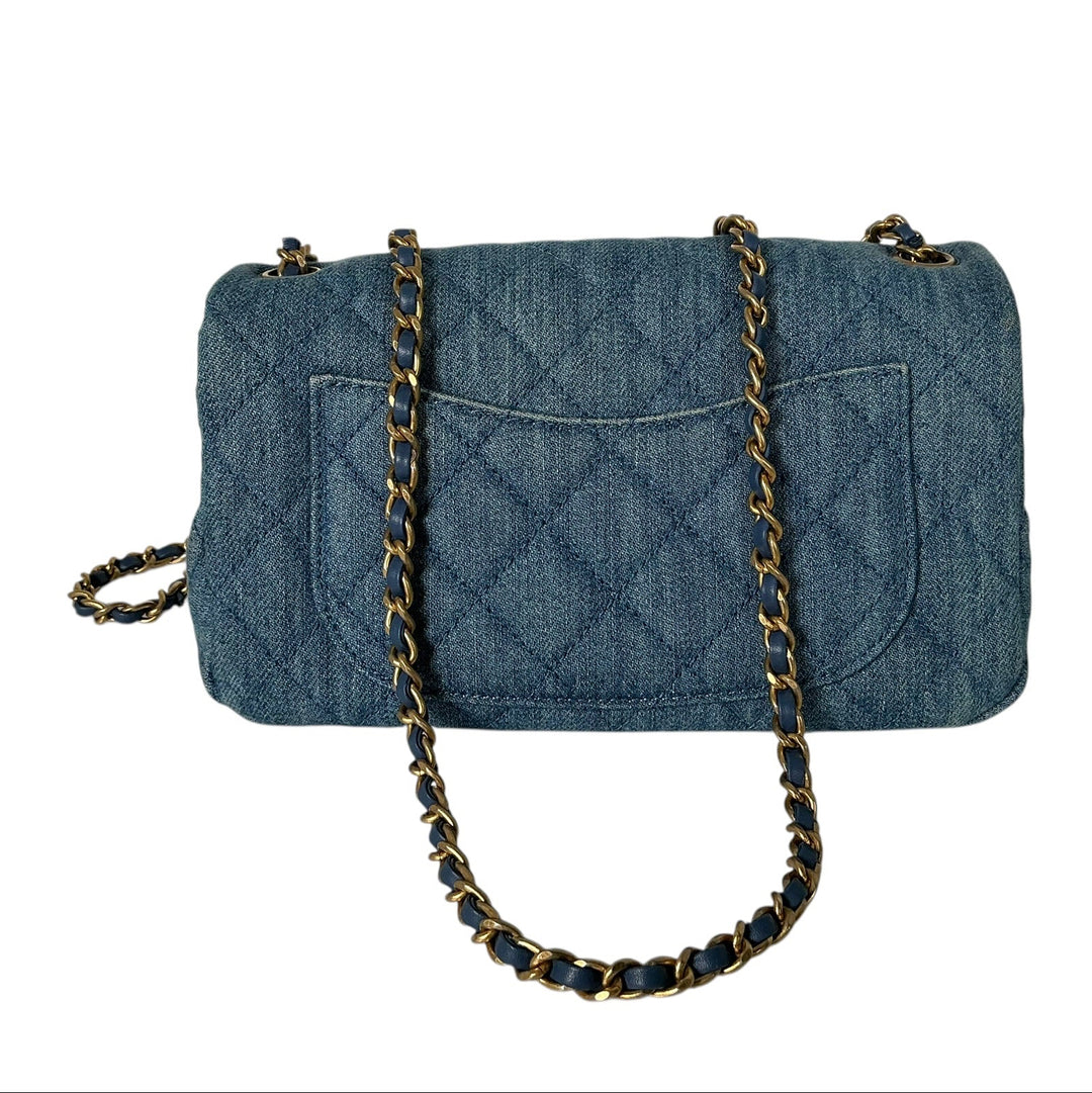 A blue quilted denim shoulder bag with a gold and black chain strap, shown against a plain white background.