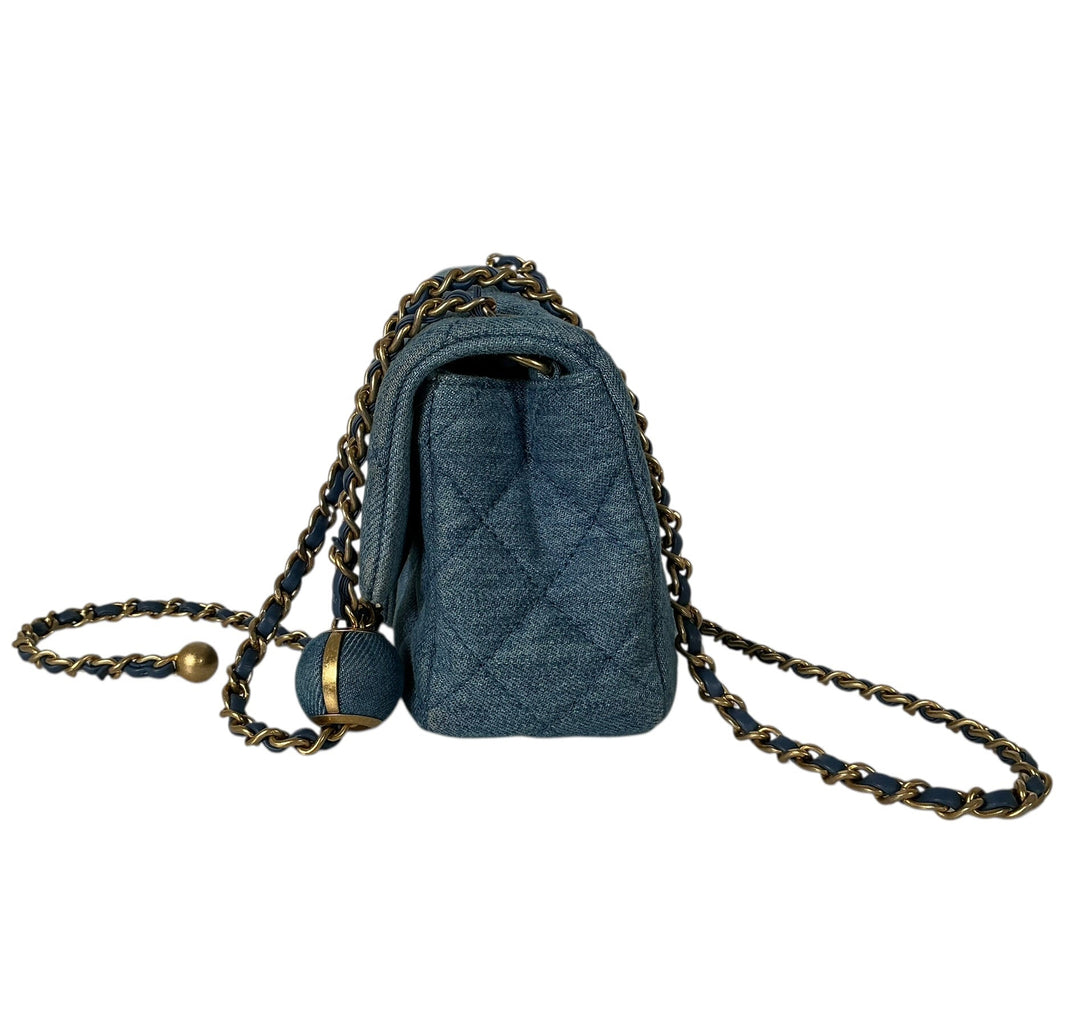 A small quilted blue shoulder bag with a gold-toned chain strap and a round blue bead charm, shown against a plain white background.