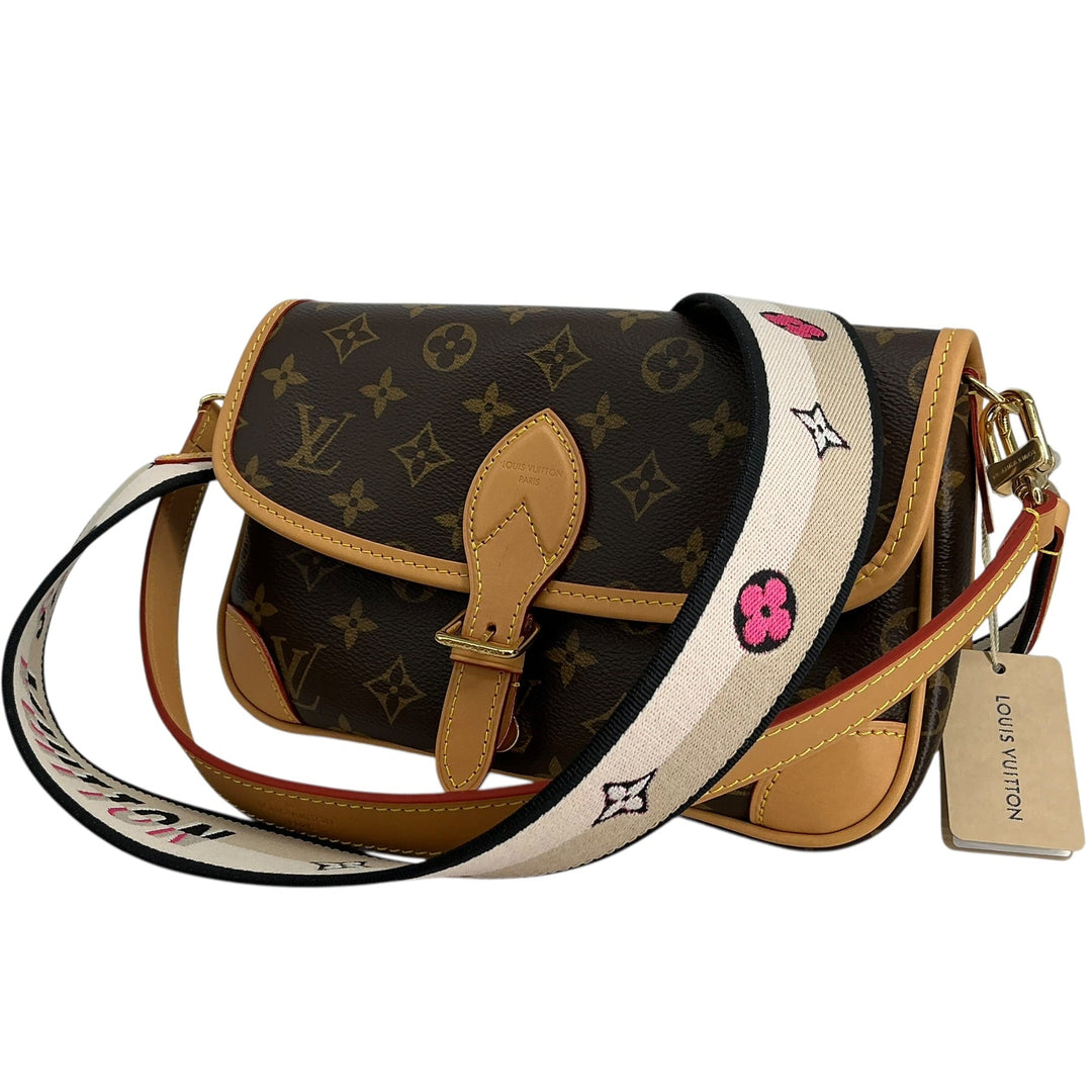 Louis Vuitton monogram canvas shoulder bag with tan leather trim and a wide white shoulder strap featuring black monogram motifs and pink flower accents, shown on a white background with a hangtag visible.