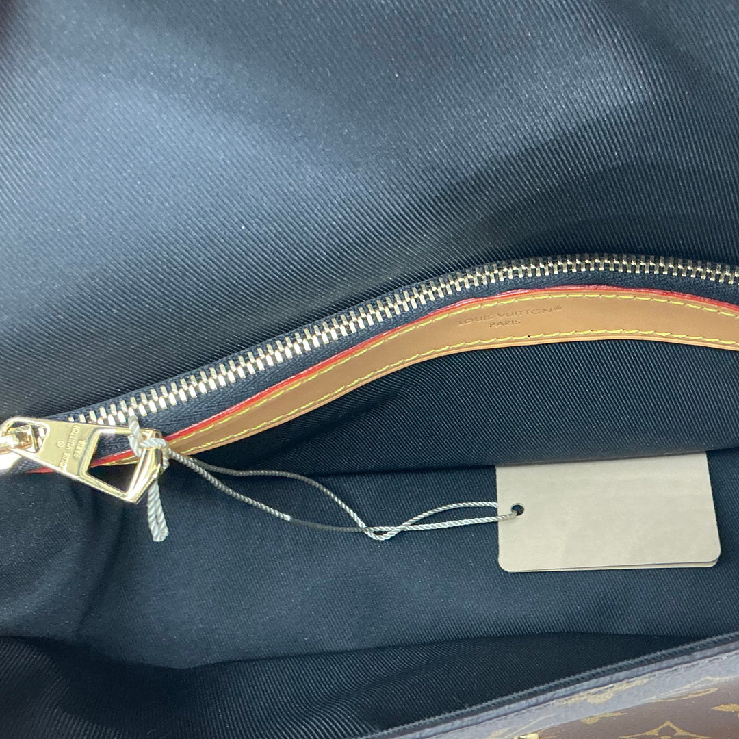 Close-up of the interior of a Louis Vuitton handbag, showing the dark navy blue fabric lining, a gold zipper pocket with a striped accent, a blank metal hangtag, and part of the brown monogram canvas and tan leather trim.