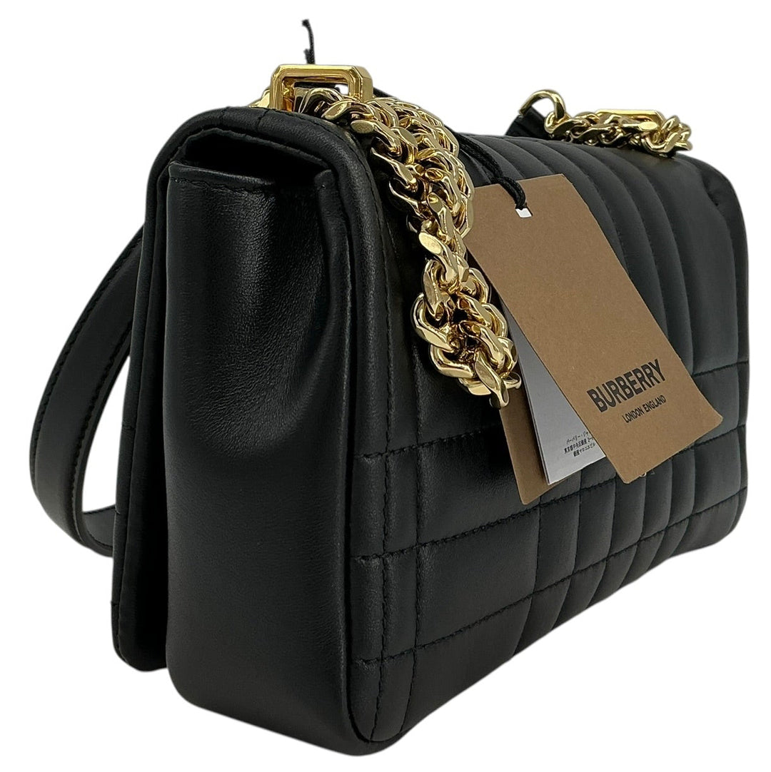 A black quilted leather shoulder bag with a gold chain strap and a brown paper Burberry hangtag, shown on a plain white background.
