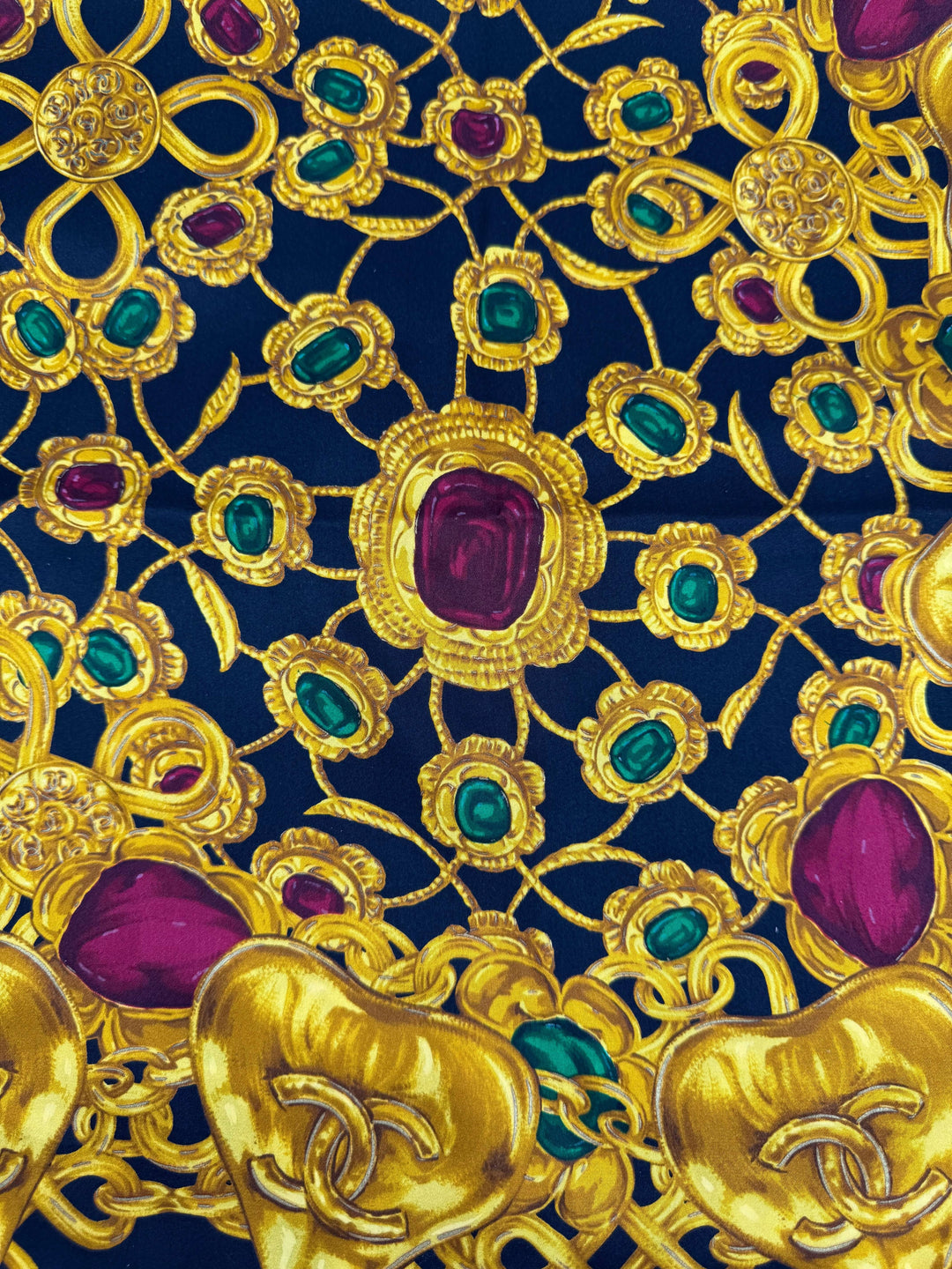 Close-up of CHANEL silk scarf with ornate gold jewelry chain design, featuring emerald green and ruby red accents on black.